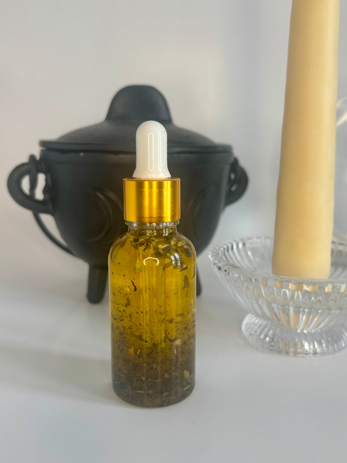 Glass bottle with gold cap and dropper filled with golden oil and herbs on a white surface with a black container and beige candle in the background.