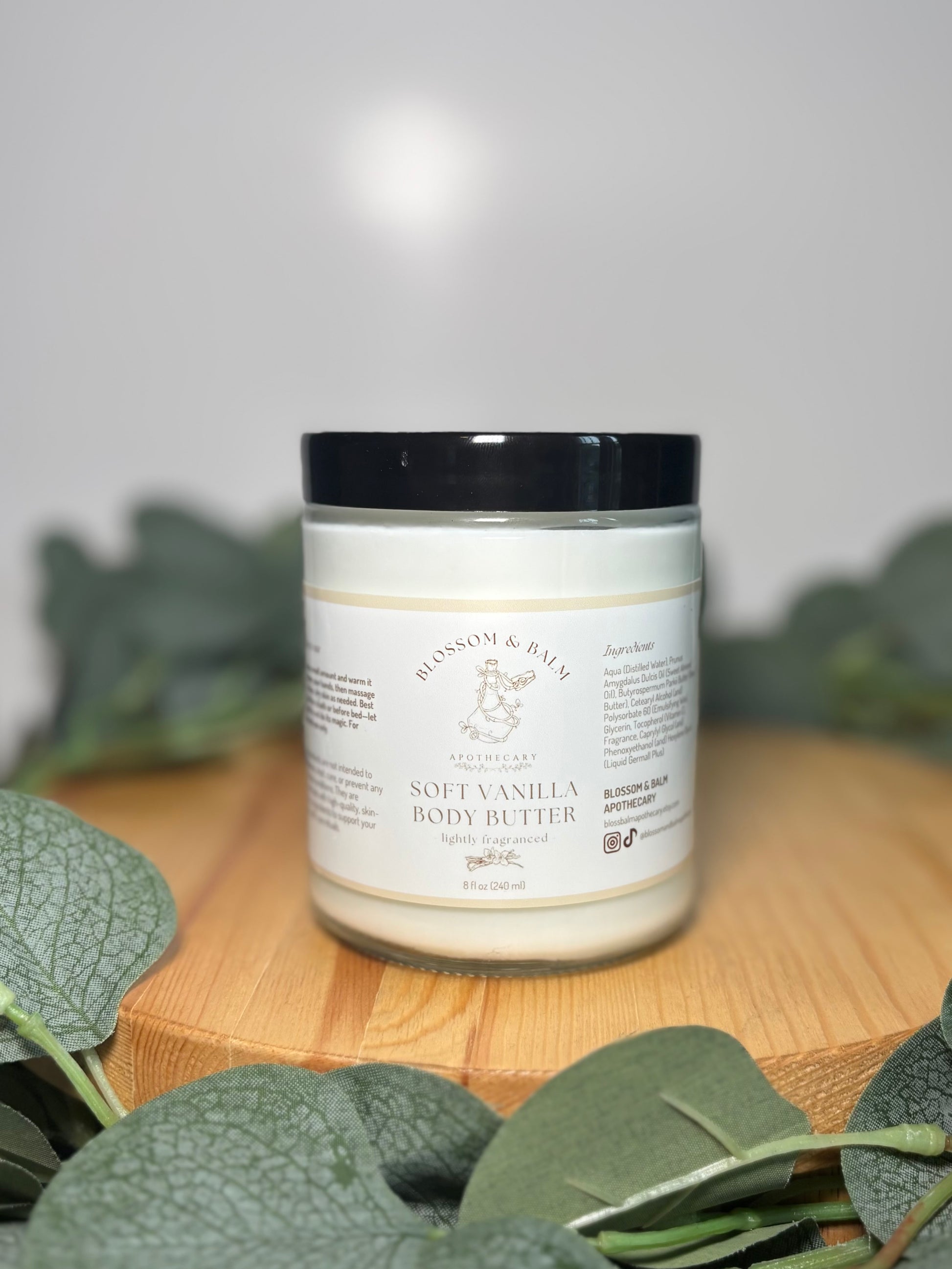 Jar of soft vanilla body butter on a wooden surface with green leaves in the background