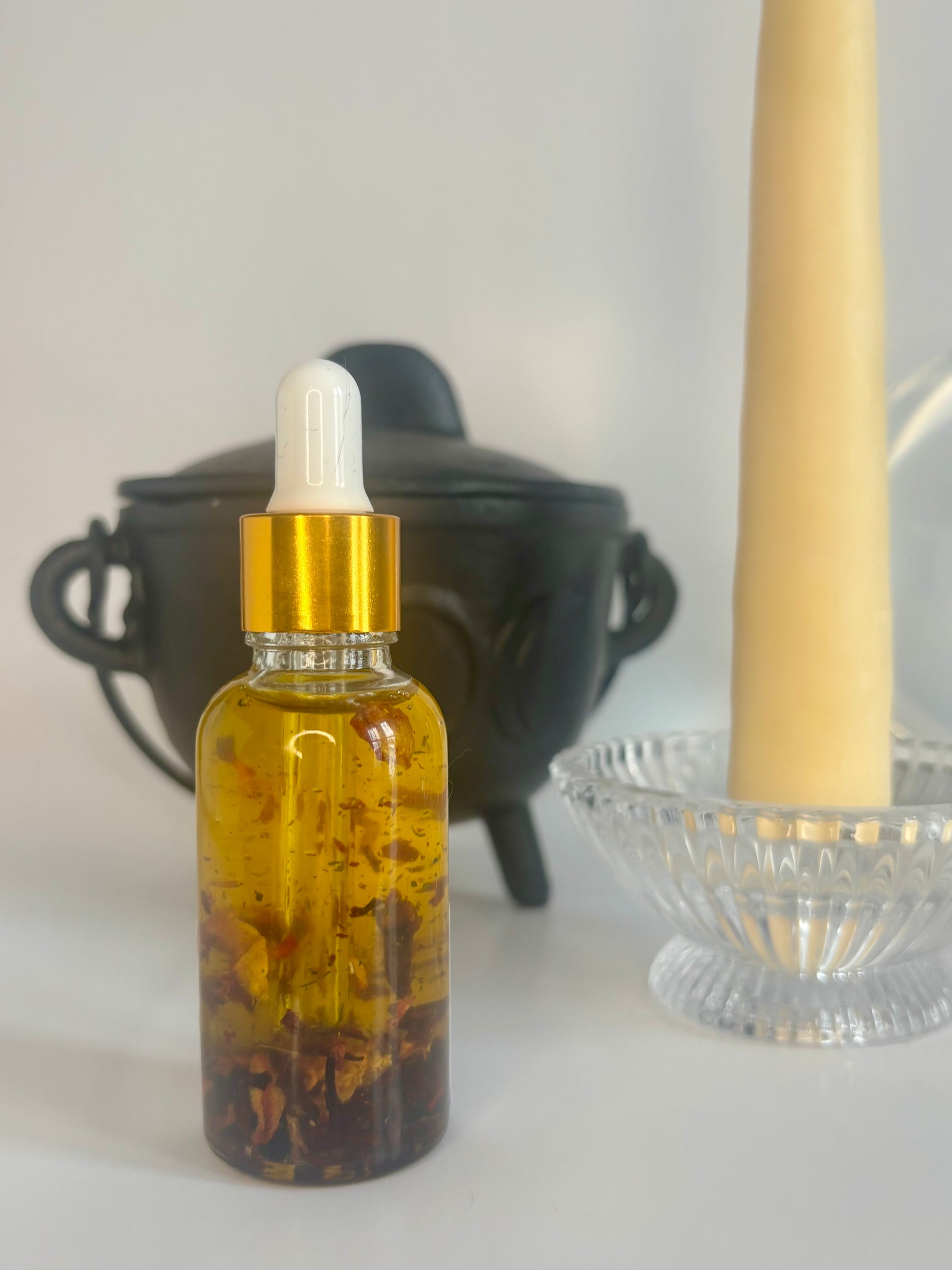 Glass dropper bottle with gold cap containing golden oil and flowers, placed on a white surface with a blurred background.