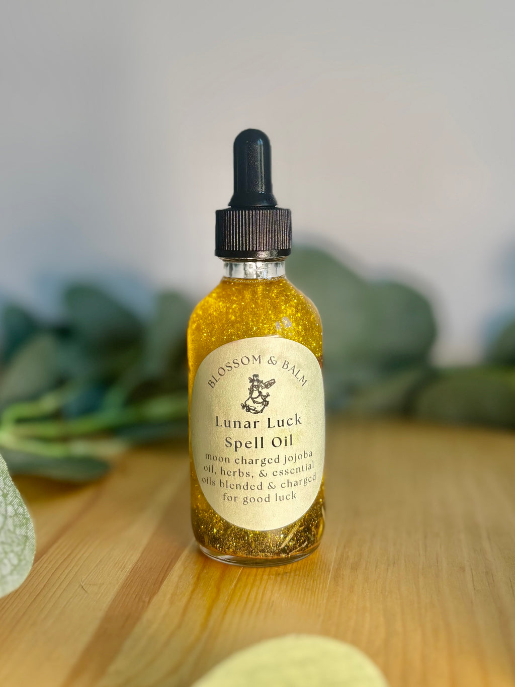 Bottle of 'Lunar Luck Spell Oil' (a bottle containing oil, herbs, and shimmering gold mica) on a wooden surface with greenery in the background.