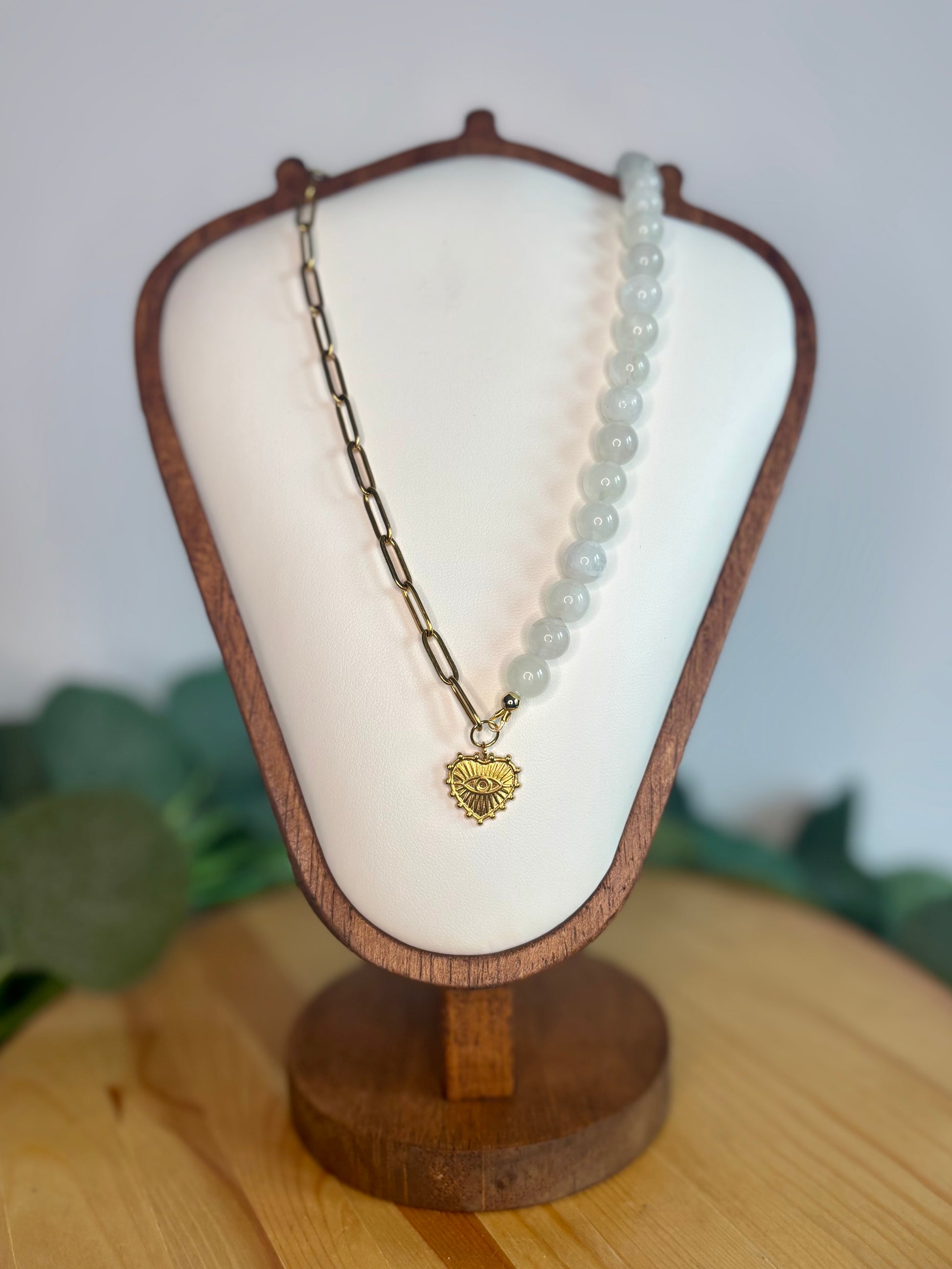 Necklace with half paperclip chain and half rainbow moonstone beads, with a heart shaped gold evil eye charm with a blurred background.