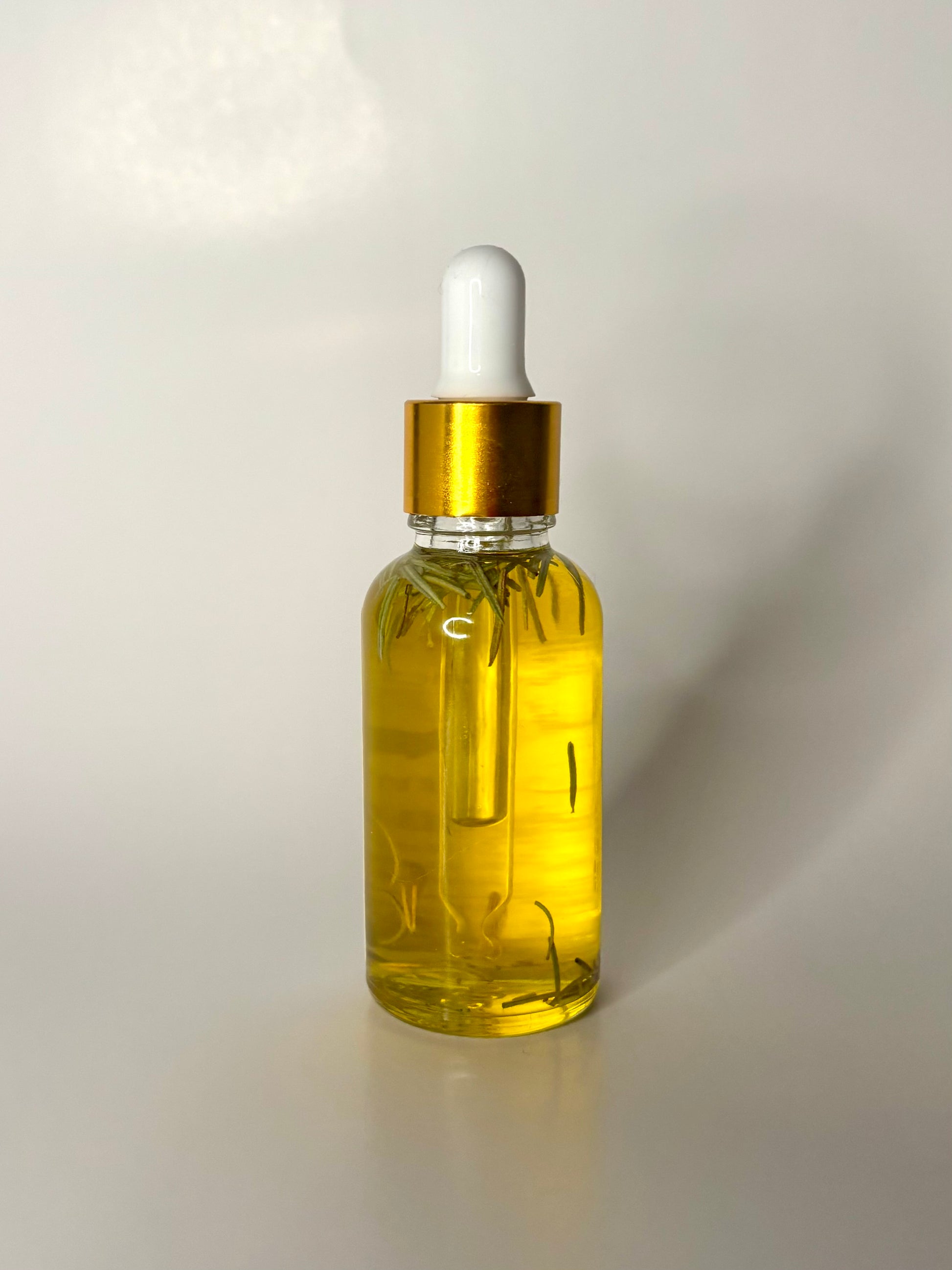 Glass dropper bottle with gold cap and white bulb filled with golden oil and floating rosemary leaves on a white background
