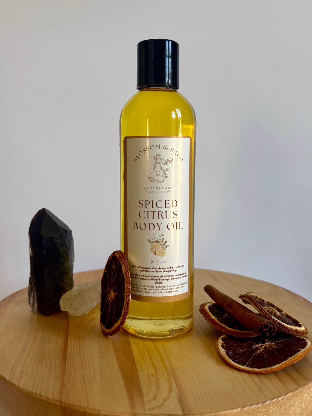 Bottle of spiced citrus body oil on a wooden surface with dried oranges and a black stone.